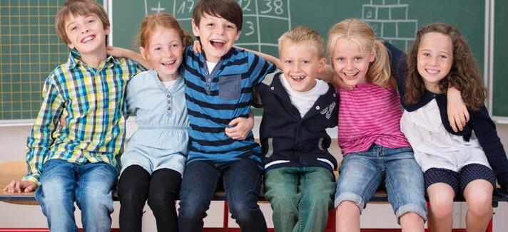 Sechs lachende Grundschulkinder sitzen vor einer Schultafel und halten sich im Arm ©contrastwerkstatt/fotolia.com